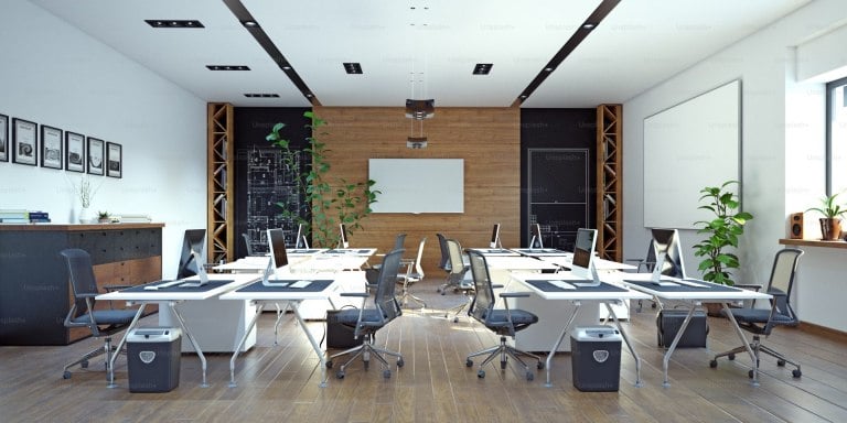 Modern office with multiple workstations, wooden accents, plants, and large windows allowing natural light. There are computer monitors on desks, chairs, and storage cabinets throughout the room.